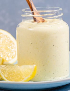 A jar of lemon pesto with a wooden spoon resting on a plate with lemon slices.