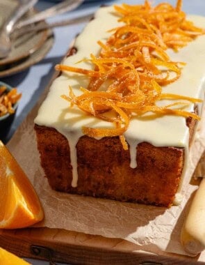 Orange cake topped with a glaze and candied orange peels sitting on a parchment lined cutting board surrounded by orange wedges and vrious other table settings.
