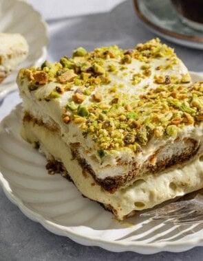 A slice of pistachio tiramisu on a plate with a fork. Nearby is another plate with pistachio tiramisu and a bowl of pistachios.