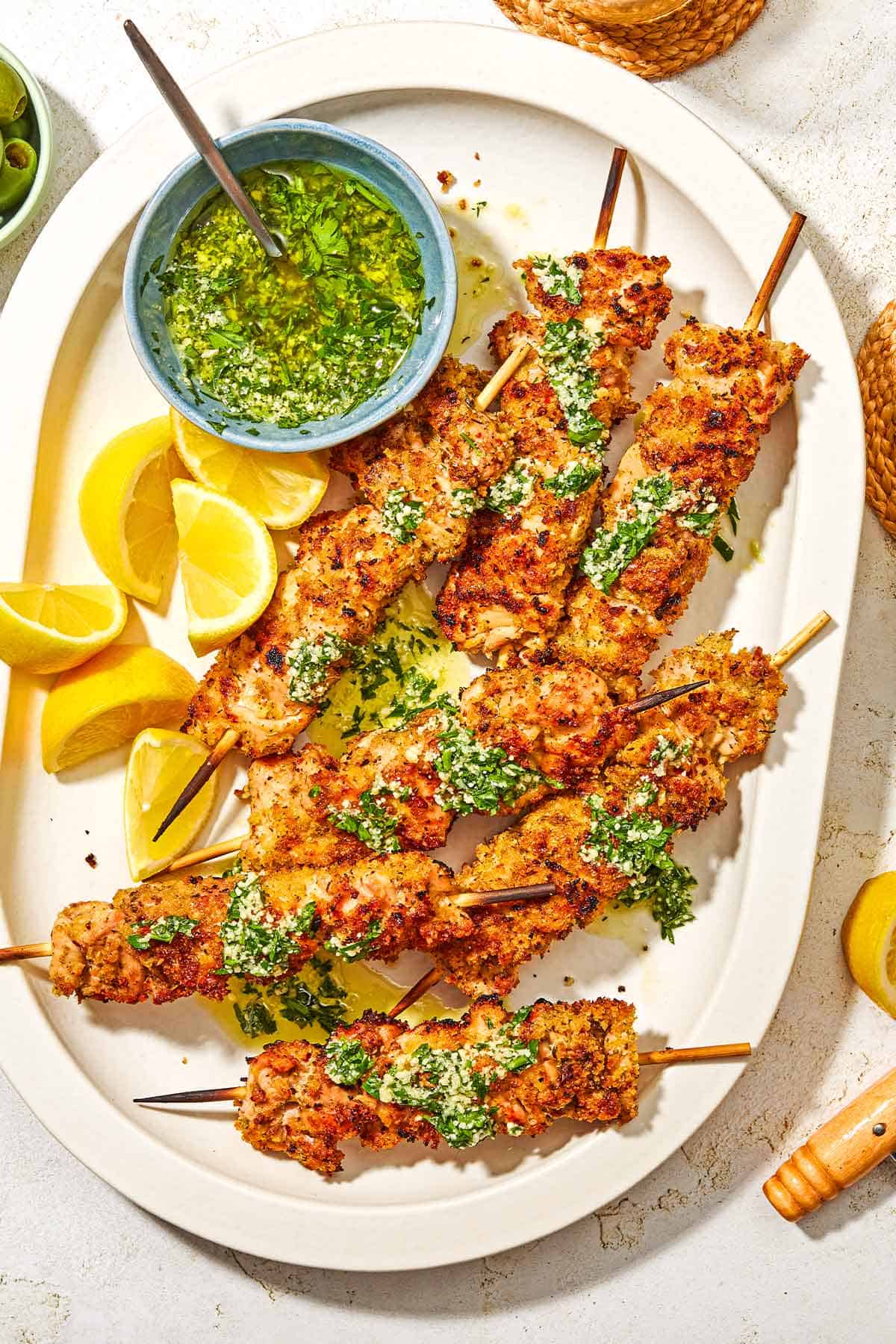 Several chicken spiedini skewers topped with garlic parmesan herb sauce in a platter with lemon wedges and a bowl of the sauce with a spoon.