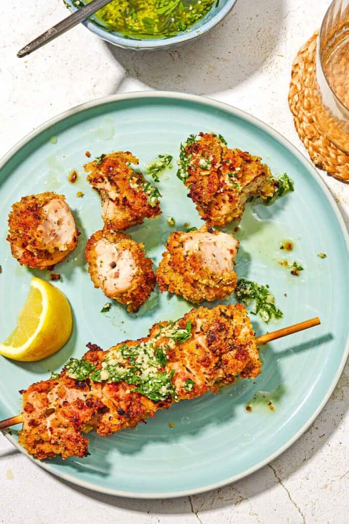 One chicken spiedini skewer on a plate along with more pieces of the chicken and a lemon wedge. Next to this is a bowl of the garlic parmesan herb sauce and a glass of wine.