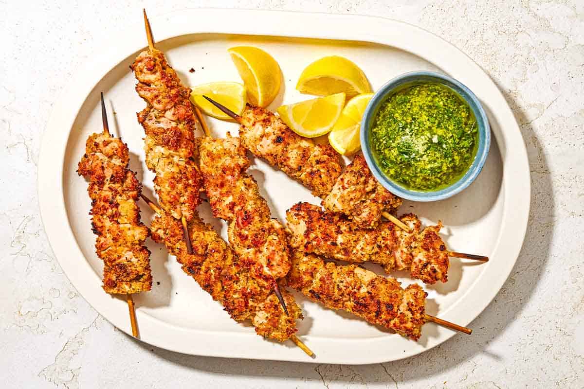 Several chicken spiedini skewers topped with garlic parmesan herb sauce in a platter with lemon wedges and a bowl of the sauce with a spoon.