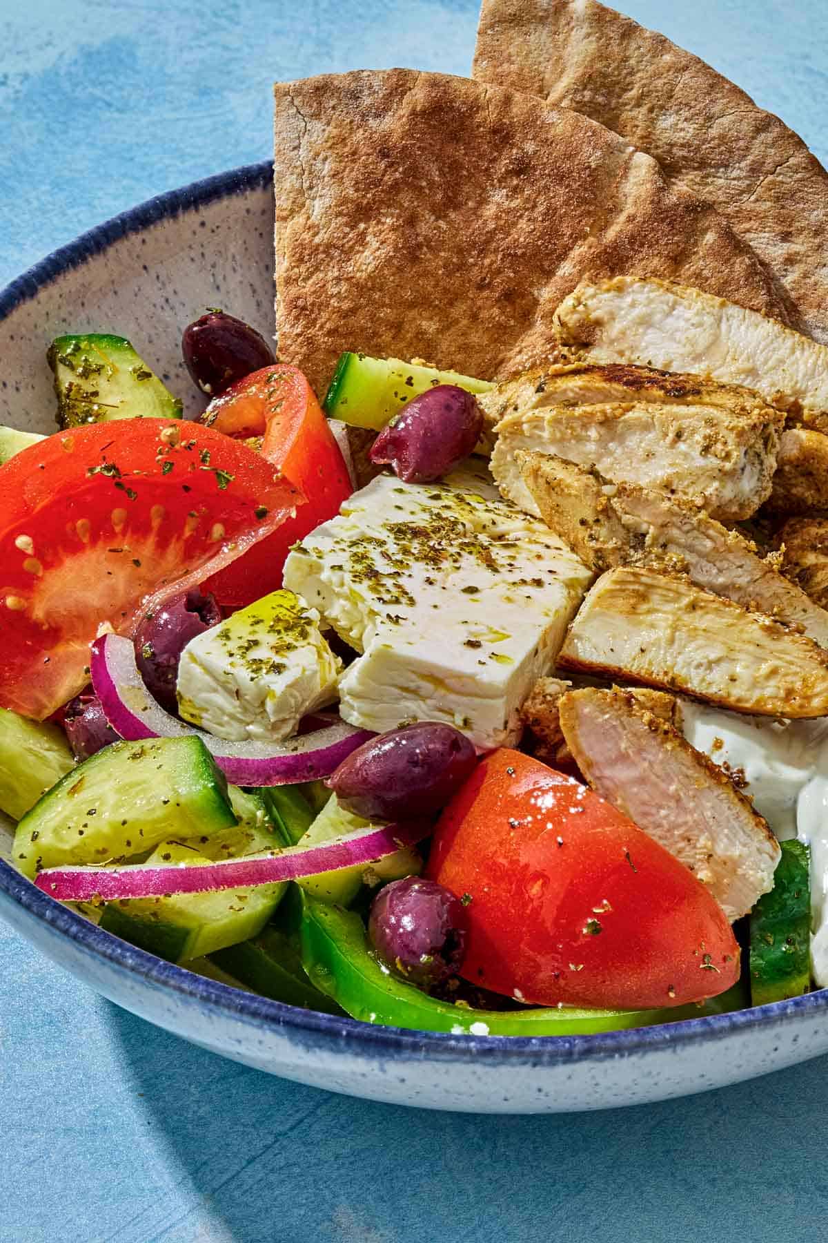 A close up photo of a gyro bowl with pita wedges, tomatoes, kalamata olives, chicken and feta.