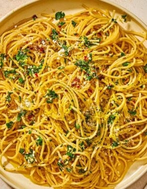 Lemon pasta topped with parsley, lemon zest, red pepper flakes and parmesan on a plate next to a fork and small bowls of red pepper flakes and parmesan cheese.