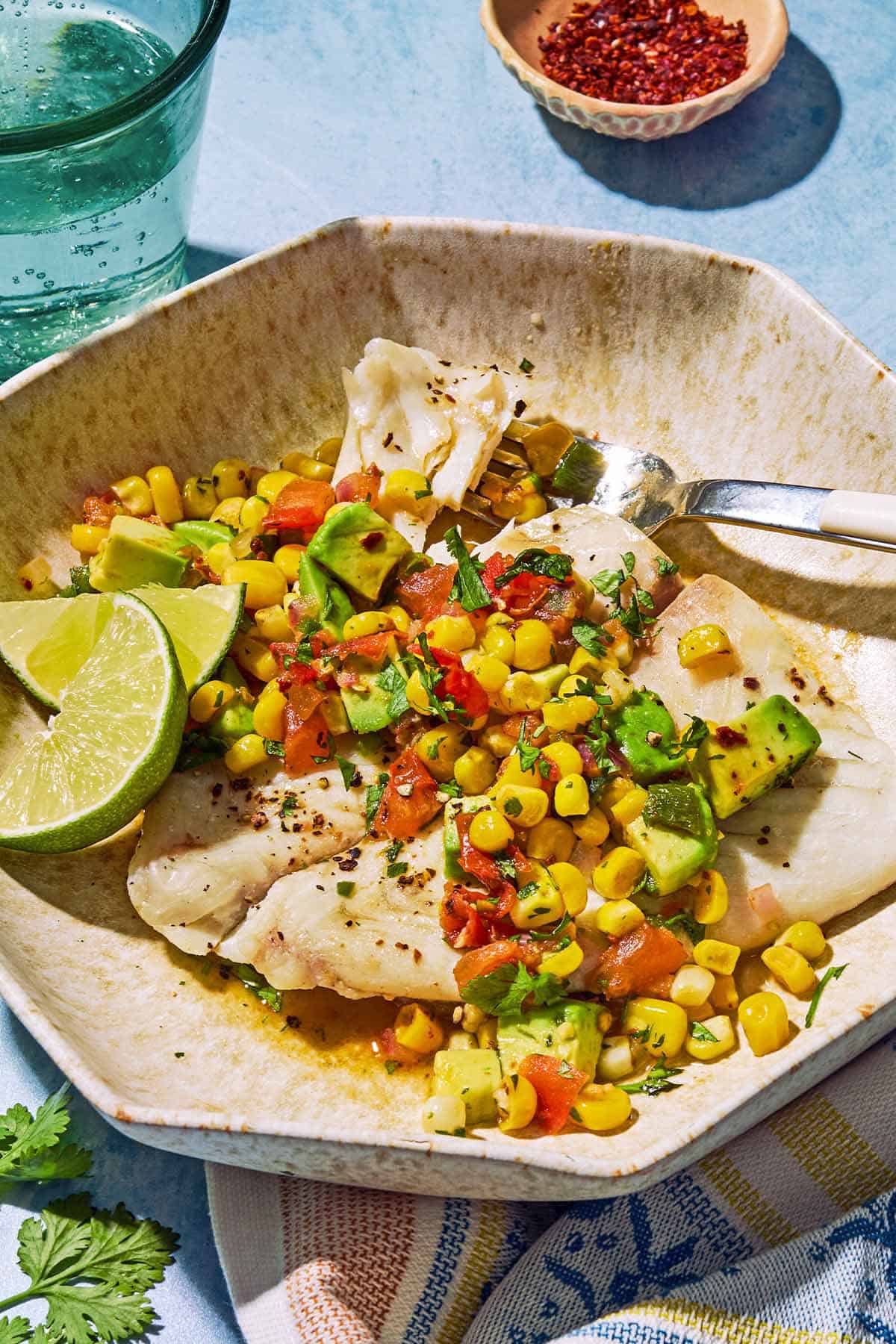 Poached flounder topped with sweet corn salsa on a plate with lime wedges and a fork. Next to this is a kitchen towel, a bowl of aleppo pepper and a glass of sparkling water.