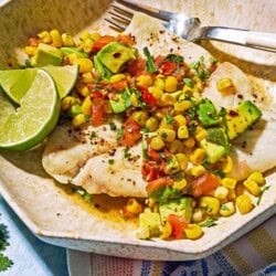 Poached flounder topped with sweet corn salsa on a plate with lime wedges and a fork.