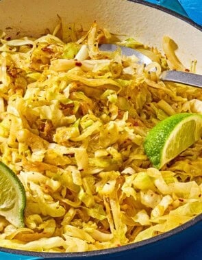 Sauteed cabbage in a skillet with a spoon and lime wedges.