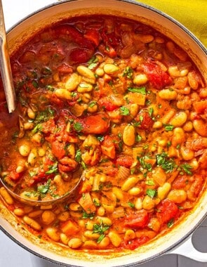 White bean stew in a large pot with a ladle next to a cloth napkin and a bowl of chopped parsley.