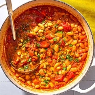 White bean stew in a large pot with a ladle next to a cloth napkin and a bowl of chopped parsley.